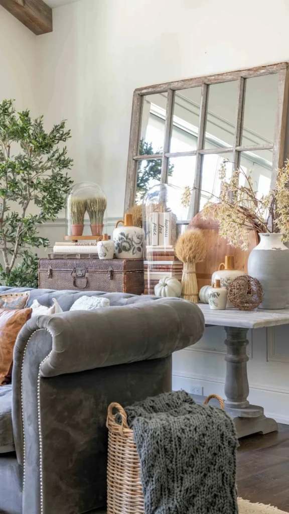 Rustic living room showcasing fall home decor ideas with a gray tufted couch, windowpane mirror, glass cloches with dried wheat, and a white pumpkin centerpiece.