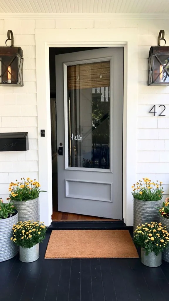 Charming cottage style front porch decor featuring a grey glass-paneled door, black vintage lanterns, and yellow flowers in galvanized metal buckets.
