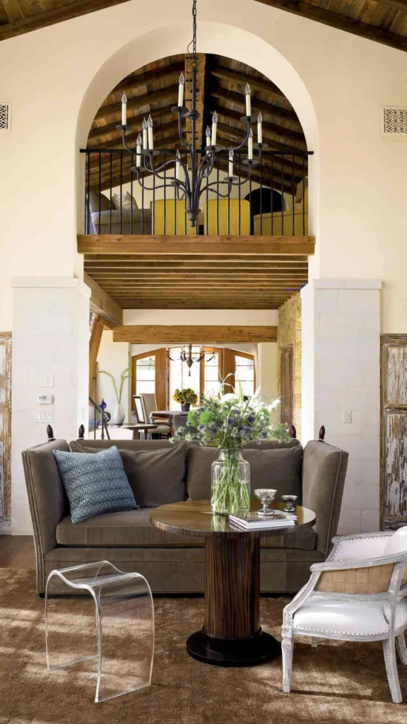 Elegant vaulted sunroom with rustic home decor featuring a high back sofa, a round wooden table, and a classic white armchair under an archway.