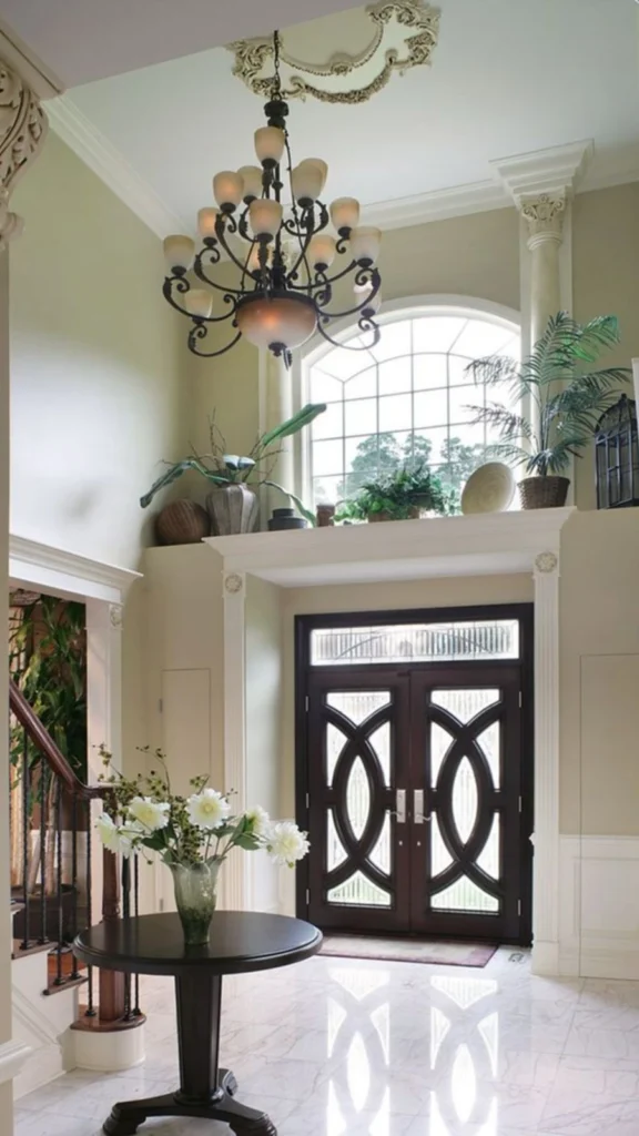 Elegant luxury home decor in a high-ceiling foyer featuring a large classic chandelier, a dark round center table with white flowers, and double entry doors with geometric glass panes.