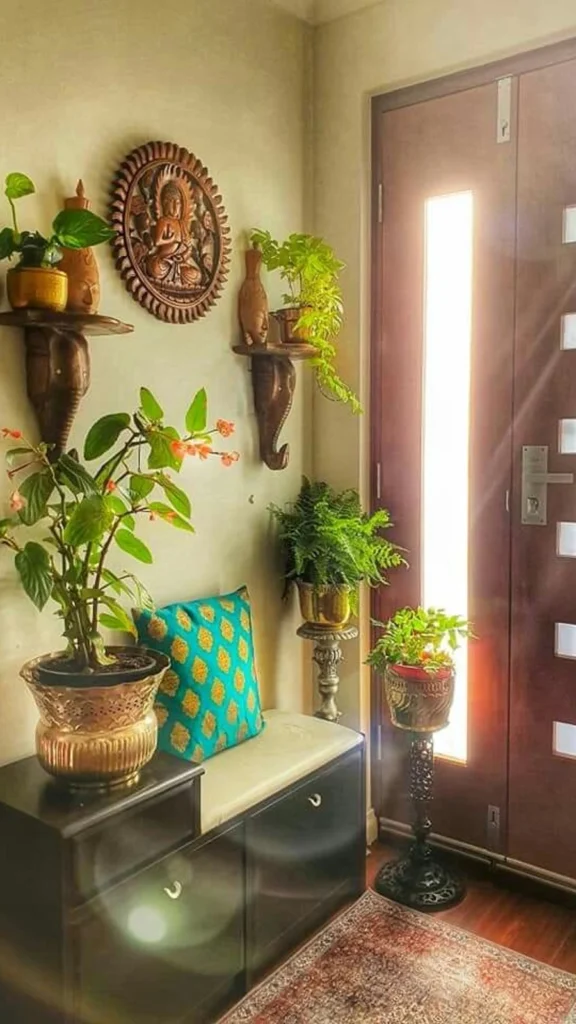 Vibrant traditional Indian entryway decor featuring indoor green plants, a wooden Buddha wall hanging, a metallic planter, and a turquoise patterned cushion on a dark storage bench.