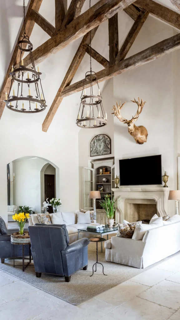 Grand double-height living room featuring rustic home decor with massive exposed wood trusses, two tiered iron chandeliers, a stone fireplace, and a mounted deer head.