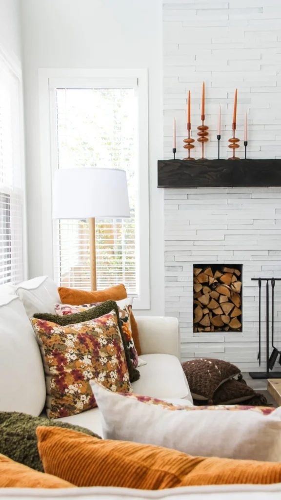 Modern living room with fall home decor ideas featuring a white brick fireplace, amber candle holders with orange taper candles, floral cushions, and stacked firewood.