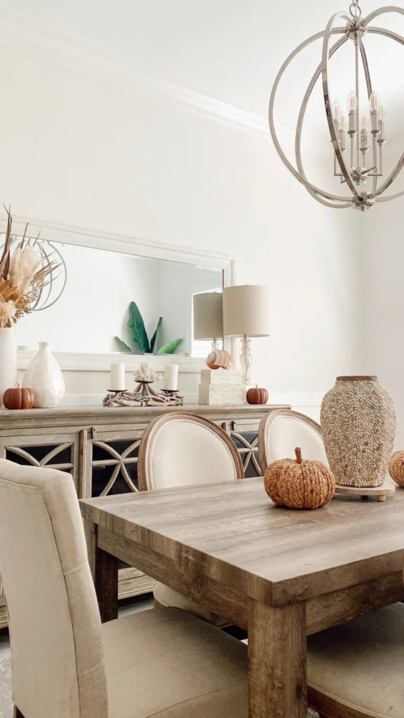Bright dining room displaying fall home decor ideas with woven decorative pumpkins on a wooden table, pampas grass, and a large wall mirror above a wooden sideboard.