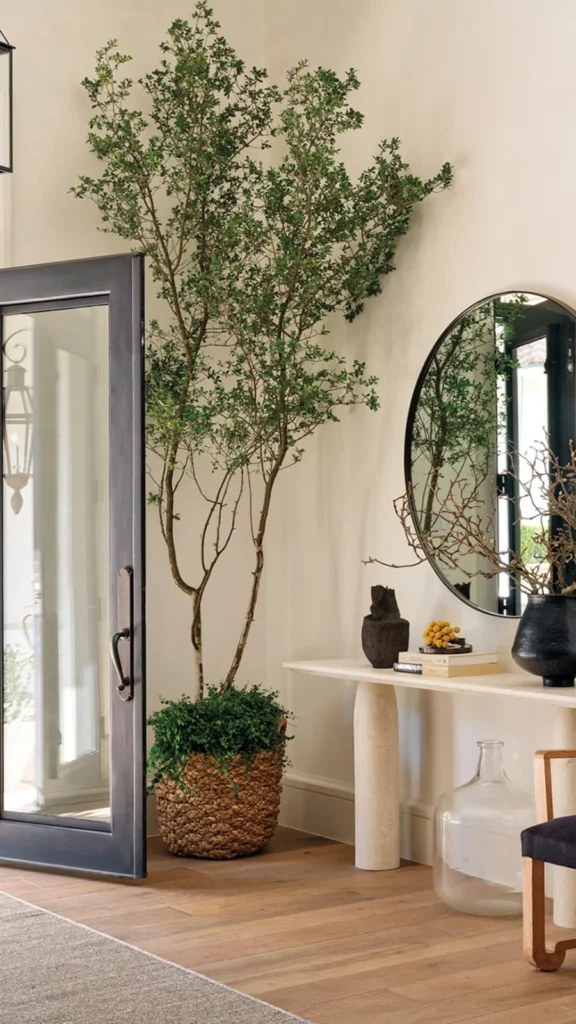 Elegant organic modern entryway featuring a tall tree in a woven basket, a sleek marble console table, a large round mirror, and a minimalist black armchair.