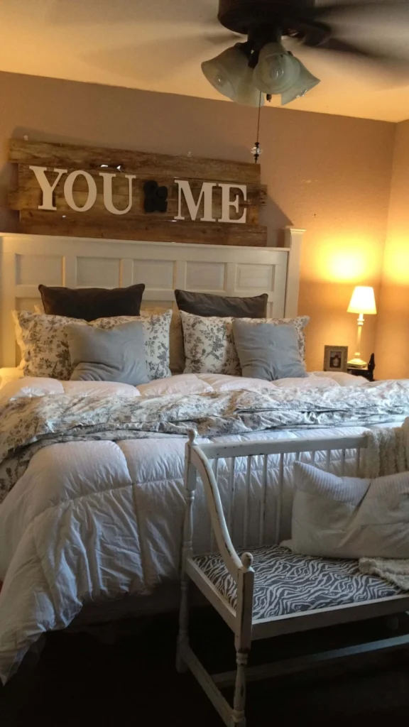 Farmhouse bedroom displaying wall decor ideas with a rustic wooden "YOU & ME" sign mounted above a white panel headboard and cozy bedding.
