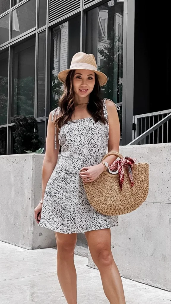 Woman wearing a white spotted tie-strap mini dress, straw sun hat, and a woven semi-circle handbag.